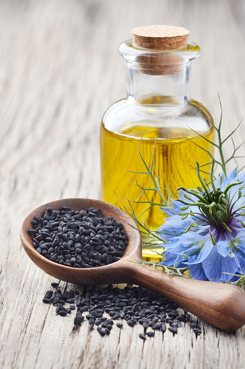 En liten flaska med svartkumminolja bredvid en svartkumminblomma och en sked svartkumminfr&ouml;n