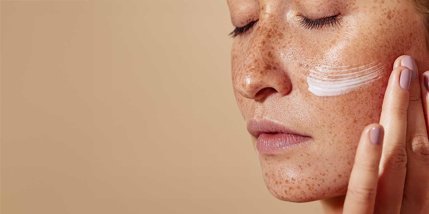 Close-up of freckled female face spreading a thin layer of cream on her cheek