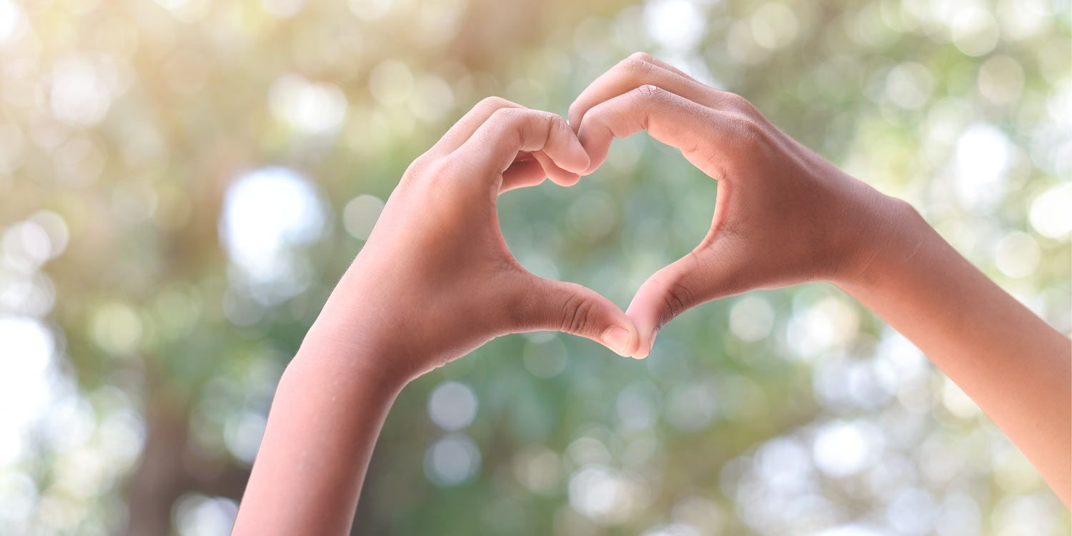Two hands held up forming a heart shape against a natural background of tree canopies