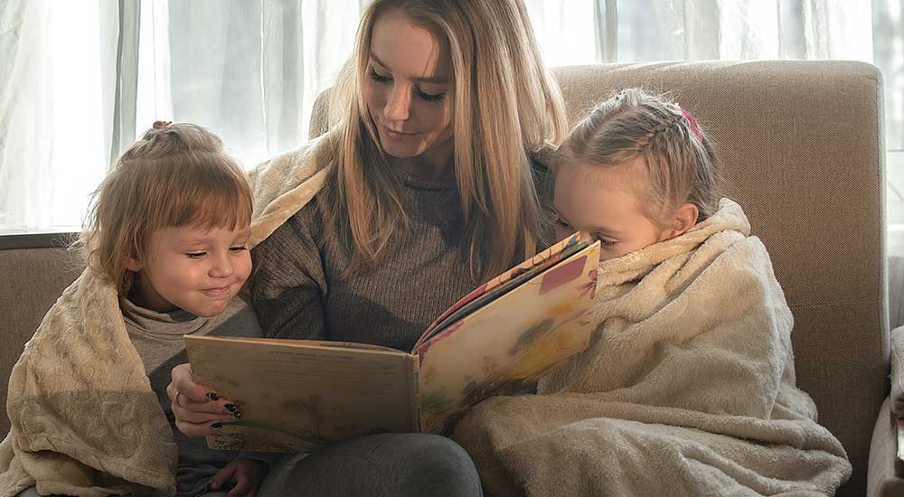 Mother reads a children's book to her two small children who are wrapped in cozy blankets