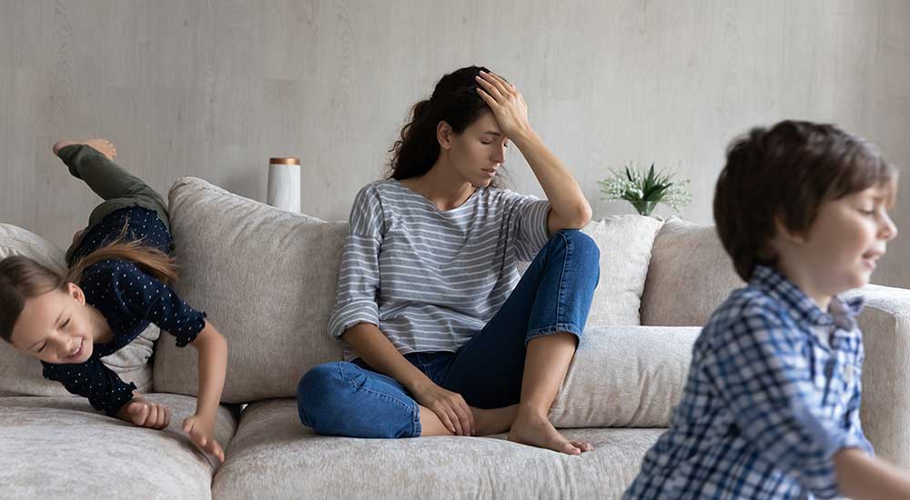 Tired mother sits on the sofa between children running around and playing