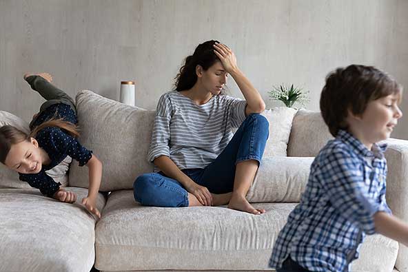 Tired mother sits on the sofa between children running around and playing