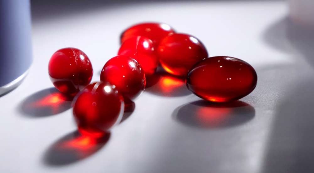 Close-up of red krill oil capsules on a white surface, lit warmly by sunlight shining through the capsules.