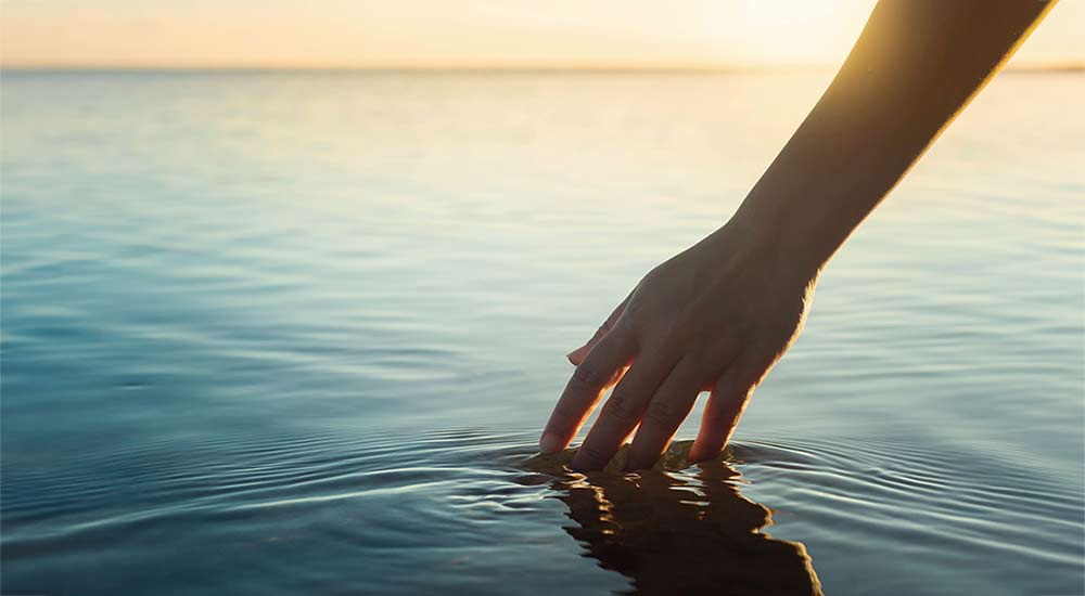 A hand touching the water surface of a lake at sunset