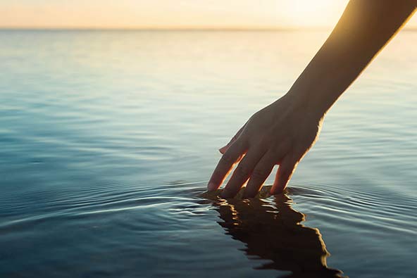 A hand touching the water surface of a lake at sunset